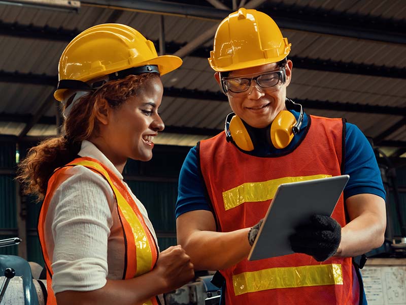 Two people wearing safety clothing in a factory, looking at a tablet device together