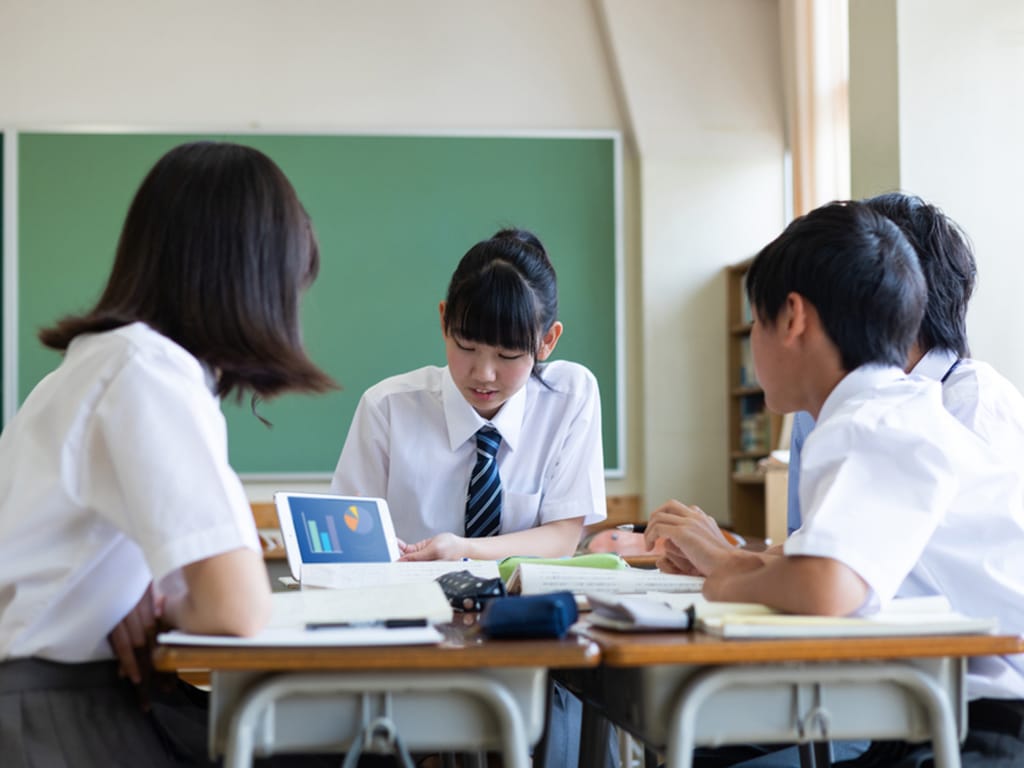 Four Japanese teenage pupils sitting at a table in a classroom with school books and a tablet device