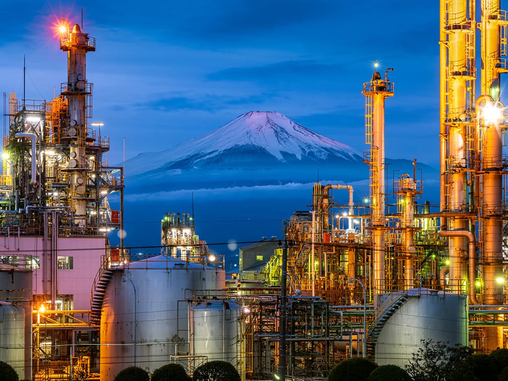 Photo of a blue-toned Mount Fuji in the background and brighly-lit industrial cranes and structures in the foreground, and at the bottom the tops of several heads looking at this image